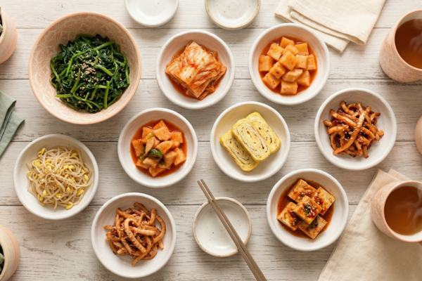 Assorted Korean banchan side dishes including spinach, kimchi, egg roll, bean sprouts, and braised tofu in small white bowls on a wooden table
